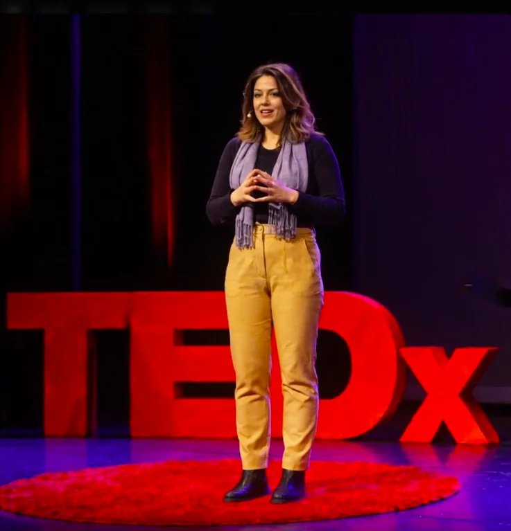 Jessica standing on a TEDx stage, full length, the TEDx letters glowing red behind her