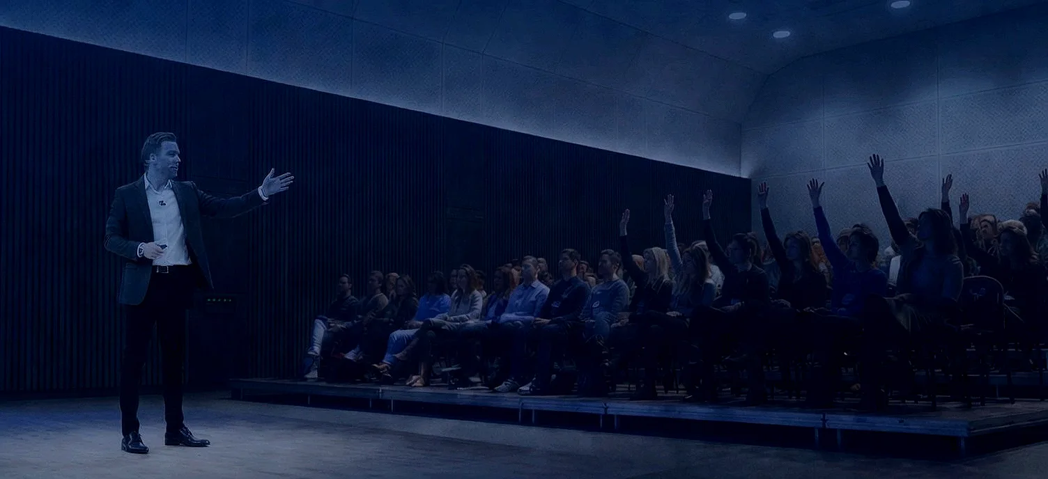 Richard on stage presenting to a dark hall with one arm out