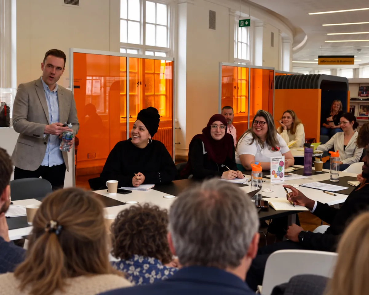 Richard standing to speak to a smiling group seated around a table in a library, notebooks and water bottles in front of them